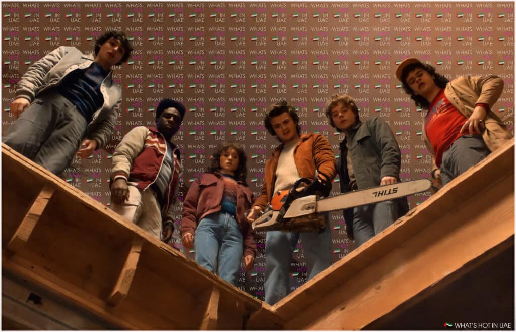 A group of six people from Stranger Things Season 5 looking down through a wooden structure, one holding a chainsaw, with a backdrop reading 'WHAT'S HOT IN UAE.'