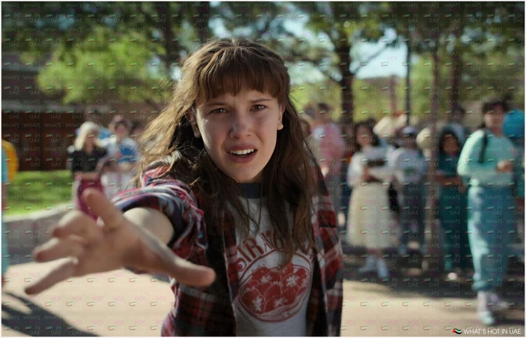 Eleven from Stranger Things Season 5 with long hair wearing a red and black checkered shirt reaches out with an intense expression, while a group of people is blurred in the background, with a subtle What's Hot in UAE wallpapered over.