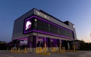 A Taco Bell restaurant with a modern design, illuminated by purple lighting, featuring a large logo on the exterior, and a drive-thru area with yellow bollards outside, set against a twilight sky.