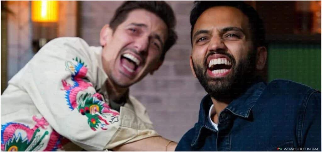 Two men laughing, one wearing a denim jacket and the other in a shirt with colorful dragon designs, in an indoor setting with warm lighting.