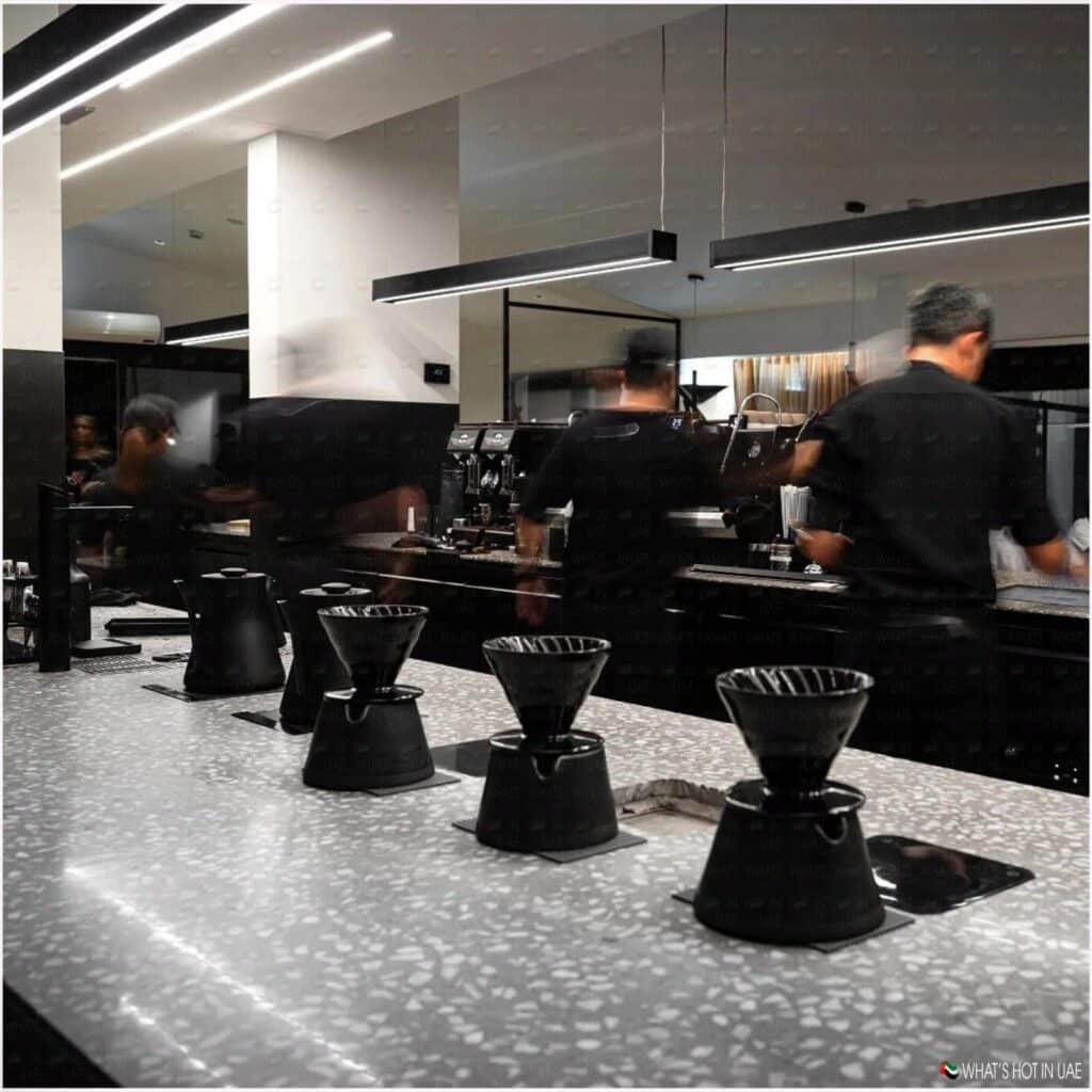 A modern coffee shop interior featuring a sleek counter with multiple black pour-over coffee brewers. In the background, baristas are seen preparing drinks at an espresso machine under bright overhead lighting.