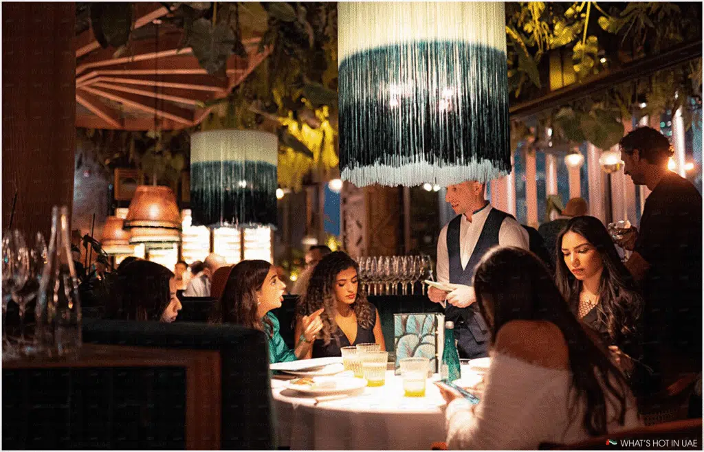 A group of people seated at a dimly lit restaurant table, with a waiter standing beside them taking orders. The table is decorated with drinks and menu items, and a fringed light fixture hangs above, contributing to the restaurant's ambiance.