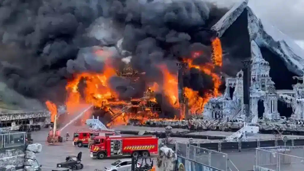 A large fire engulfs a snowy-themed structure, with bright orange flames and thick black smoke billowing into the sky. Fire trucks are parked nearby, and hoses are spraying water onto the fire.