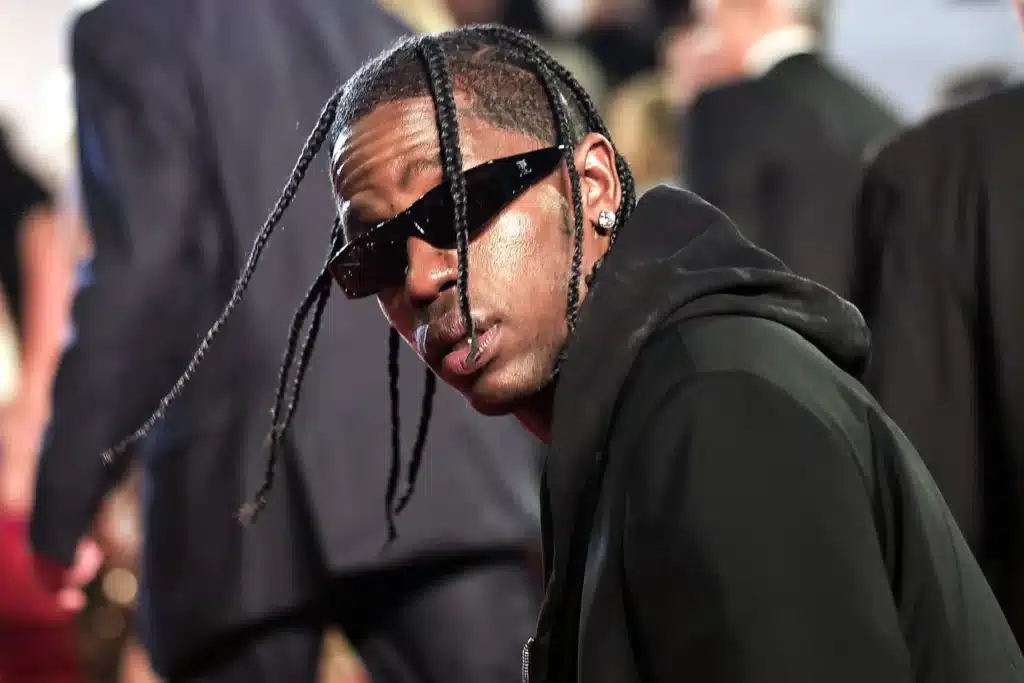 A person wearing sunglasses and a black hooded jacket, with braided hair, looking towards the camera in a crowded setting.