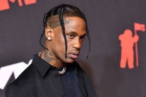 A person with braided hair and tattoos on the side of their head, wearing a black shirt and a beaded necklace, poses against a dark background with a red silhouette of a figure holding a flag.