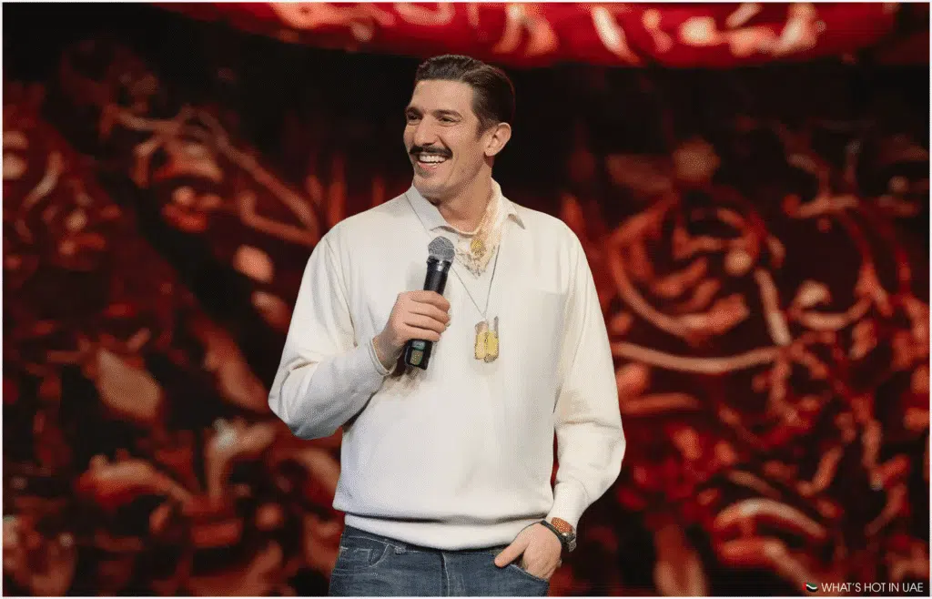 Andrew Schulz with a microphone stands on stage, wearing a cream-colored sweater and jeans, with a patterned red backdrop behind him. This represents the comedy shows within UAE Nightlife