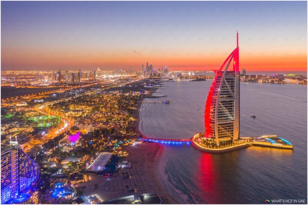 Aerial view of Dubai at twilight, featuring a brightly lit, sail-shaped building on the water's edge, with the city's illuminated skyline and a colorful sky in the background.