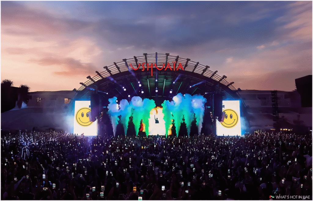A large outdoor concert stage at sunset with colorful smoke plumes and a crowd holding up phones, with "USHUAIA" illuminated above the stage.