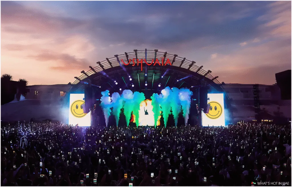 A large crowd is gathered at an outdoor concert stage at Ushuaia in Dubai with colorful smoke and light effects. The stage features a backdrop displaying a large smiley face, and the logo 'Ushuaïa' is prominently displayed at the top. Many attendees are holding up their phones to capture the event. The sky is at dusk, adding a vibrant atmosphere to the scene.
