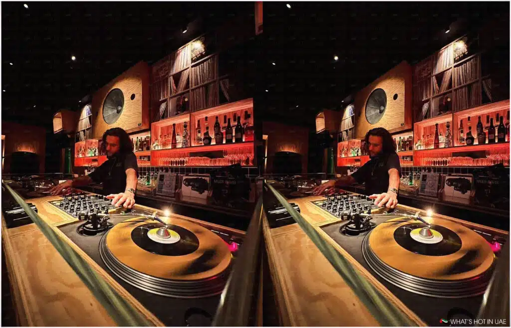 A DJ is playing music on turntables in a warmly lit bar with shelves of records and bottles in the background.