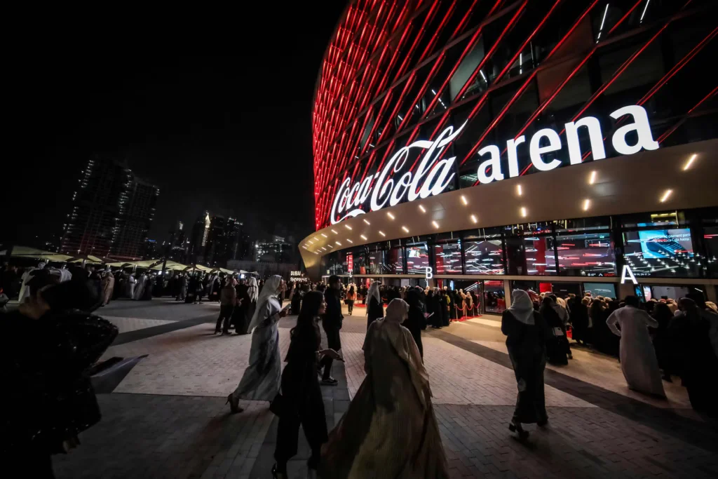 Crowd of people approaching the Coca Cola Arena at night, featuring a prominent illuminated sign and vibrant red lighting on the building, with modern skyscrapers in the background.