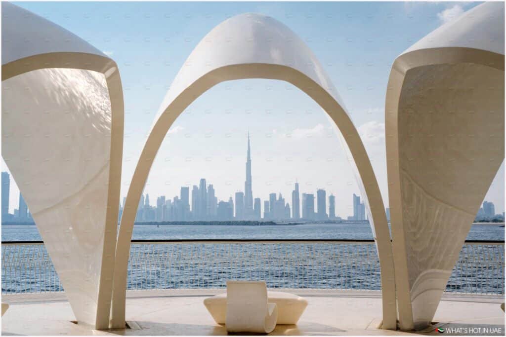 A modern architectural structure with curved arches frames a distant city skyline across a body of water on a sunny day.