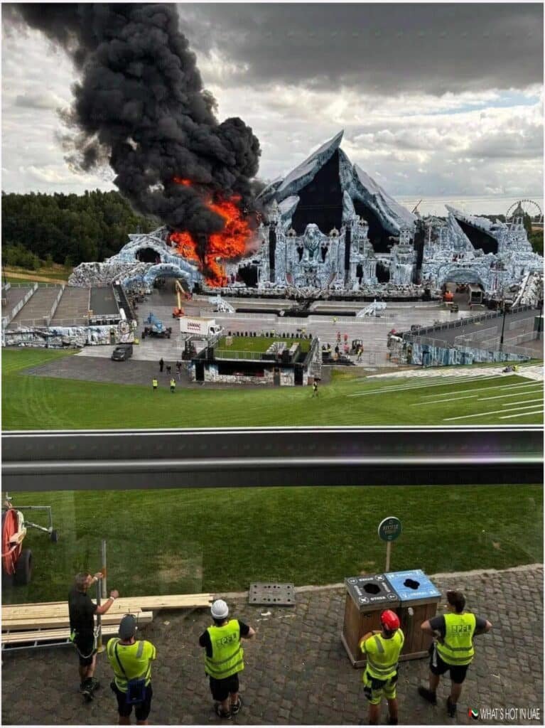 An outdoor stage designed to resemble icy mountains is engulfed in flames and thick black smoke. Below, several workers wearing safety vests watch the fire from a distance across a grassy field.