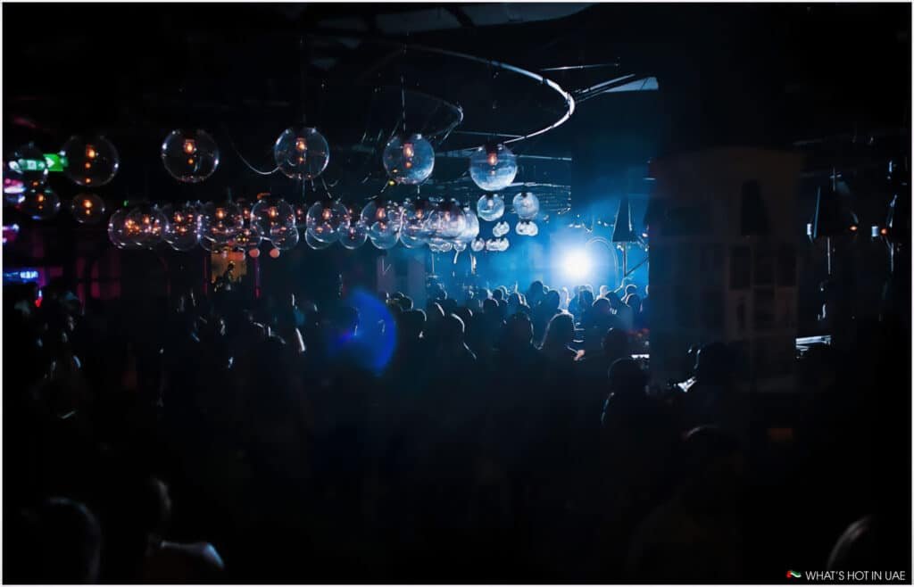A lively nightclub scene with a crowded dance floor, dim lighting, and numerous hanging spherical lights, set against a backdrop of blue and purple hues.