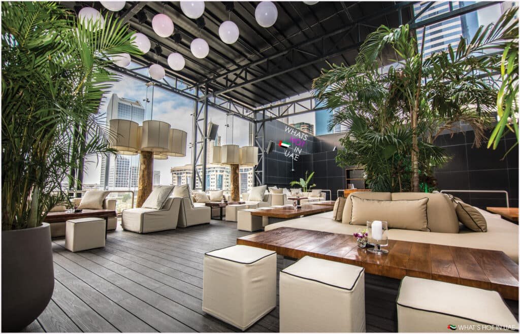 A stylish outdoor lounge with cushioned seating, wooden tables, and large potted plants, under a roof adorned with glowing spherical lamps. The city skyline is visible through large windows, and a sign on the wall reads "What's Hot in UAE."