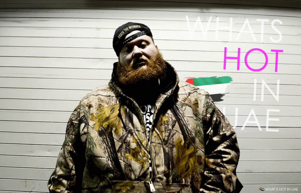 A person with a bushy red beard and wearing a camouflage jacket and a cap stands in front of a wall with text reading "What's Hot in UAE."