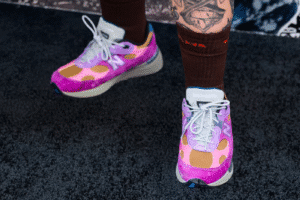 A pair of colorful sneakers with a pink, purple, and orange design, worn with brown socks and visible tattoos on the legs.