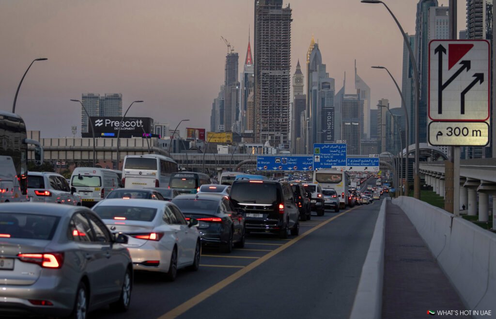 A busy highway in Dubai with heavy traffic, surrounded by skyscrapers and modern buildings in the background, including visible signs and a traffic sign indicating a junction in 300 meters.
