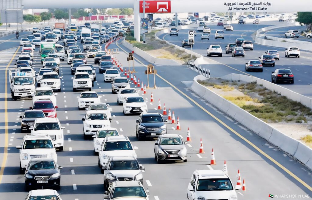 A busy multi-lane highway with dense traffic traveling in both directions near the Al Mamzar Toll Gate. Orange cones separate some lanes, and there are various types of vehicles, including cars and small trucks, under a clear sky.