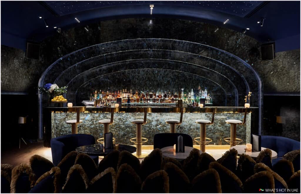 Luxurious bar interior with a curved, illuminated counter and high stools, set against a dark, reflective backdrop with various bottles displayed; plush seating in the foreground.