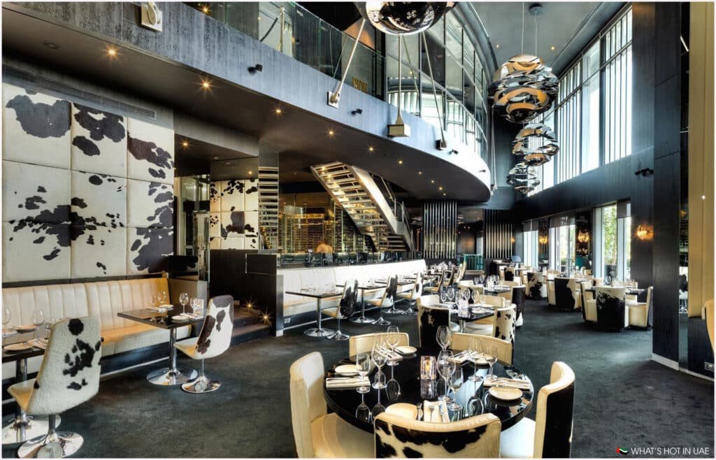 Modern restaurant interior with cow-print upholstered chairs and booths, glossy black tables, and large spherical hanging light fixtures in a spacious setting with high glass walls.