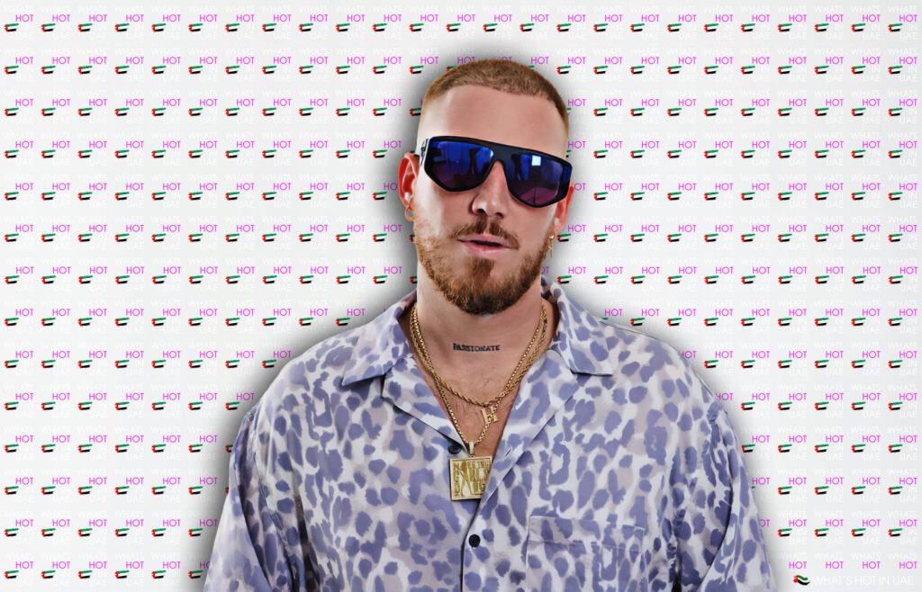 A man wearing sunglasses, a leopard-print shirt, and layered gold necklaces stands in front of a backdrop featuring the words 'WHAT'S HOT IN UAE' along with small UAE flags. Hugel Live at Pure Pacha