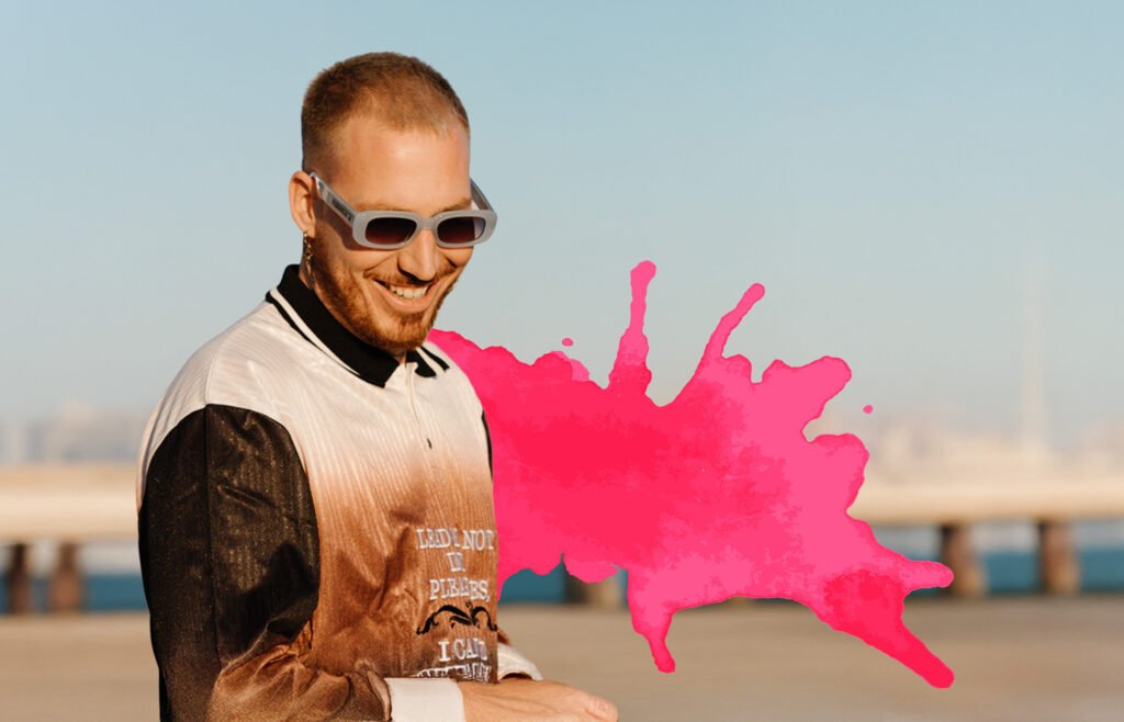 A person outdoors wearing sunglasses and a multicolored shirt, smiling with a bright pink paint splatter effect beside them.