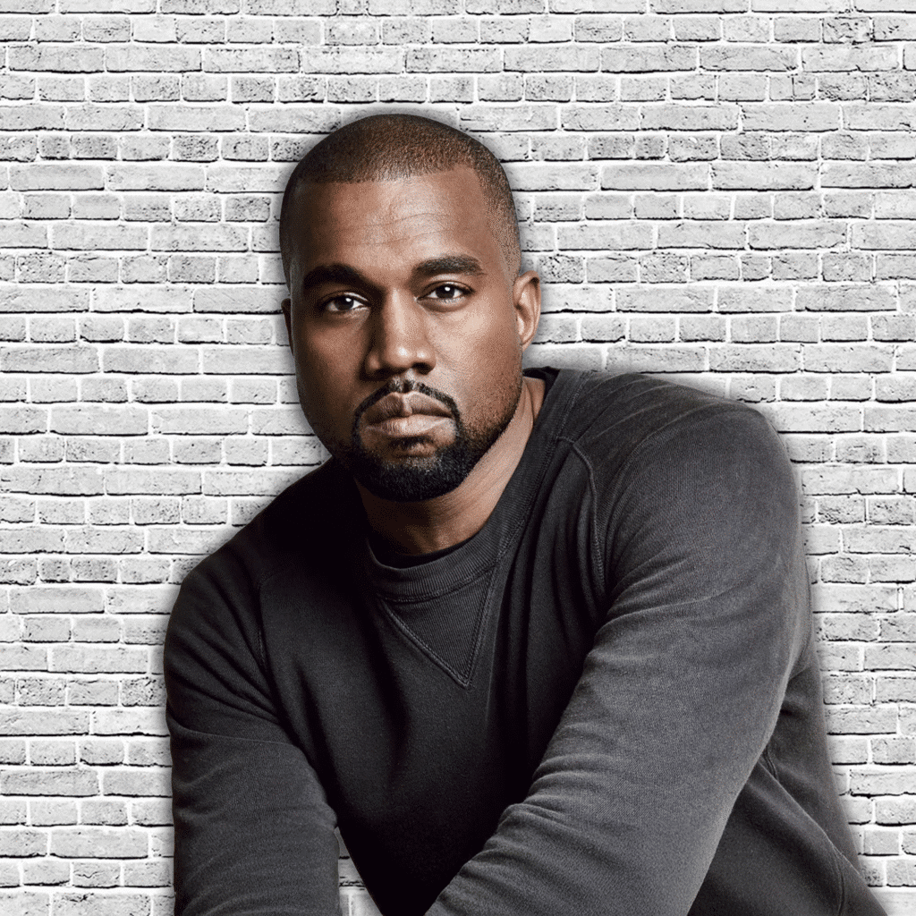 A man in a dark sweater stands in front of a white brick wall, arms crossed and gazing forward.