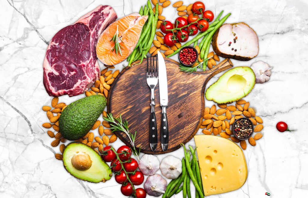 A variety of foods including raw steak, salmon, green beans, cherry tomatoes, sliced chicken, whole and sliced avocado, sliced cheese, almonds, and garlic arranged around a wooden cutting board with a knife and fork on it, all placed on a marble surface.