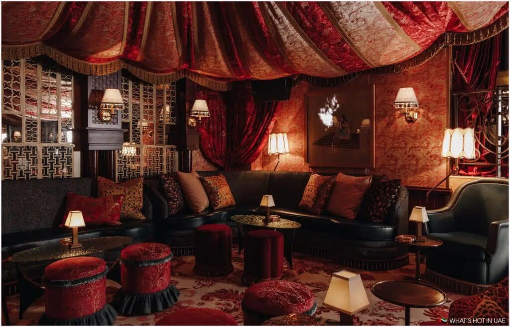 Luxurious lounge interior with red and gold draped ceiling, dark upholstered seating, patterned pillows, and warm lighting from table lamps and wall sconces, creating an opulent and cozy ambiance.