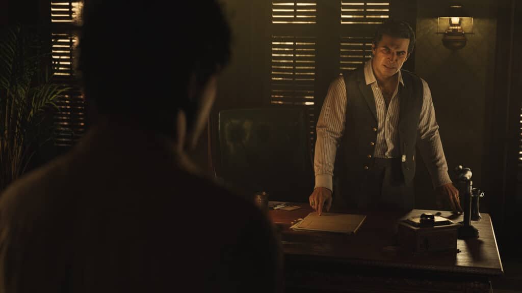 A person standing behind a desk in a dimly lit room, wearing a striped shirt and vest, with slatted window blinds providing a soft, moody backlight. Upcoming new game releases.