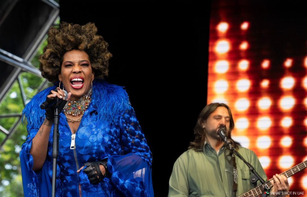 Macy Gray live in Dubai in a vibrant blue outfit singing passionately into a microphone, with another musician playing guitar in the background against an illuminated backdrop.