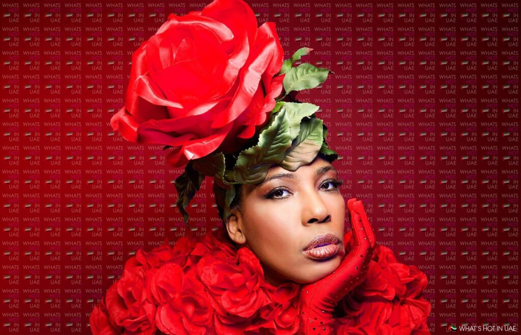 Person wearing a large red rose hat and a red floral outfit against a background with 'WHAT'S HOT IN UAE' text repeated. The individual rests their head on a gloved hand, looking at the camera.