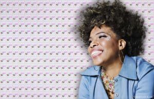 A smiling woman with curly hair, wearing a blue jacket and large hoop earrings, set against a background pattern of "WHAT'S HOT IN UAE" text and UAE flags.