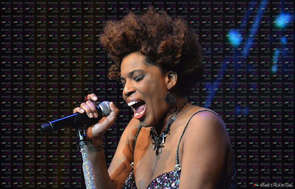 Macy Gray live in Dubai with an afro hairstyle passionately singing into a microphone on stage, wearing sparkly attire and large earrings against a backdrop labeled 'What's Hot In UAE.'