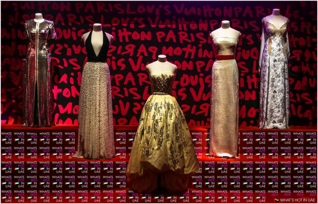 A display of five elegant Louis Vuitton gowns on mannequins against a background with "Louis Vuitton Paris" written in bold red letters. The gowns are made from various materials, featuring metallic and beaded embellishments, with styles ranging from strapless to halter neck designs. The floor features a pattern of the text "WHAT'S HOT IN UAE."