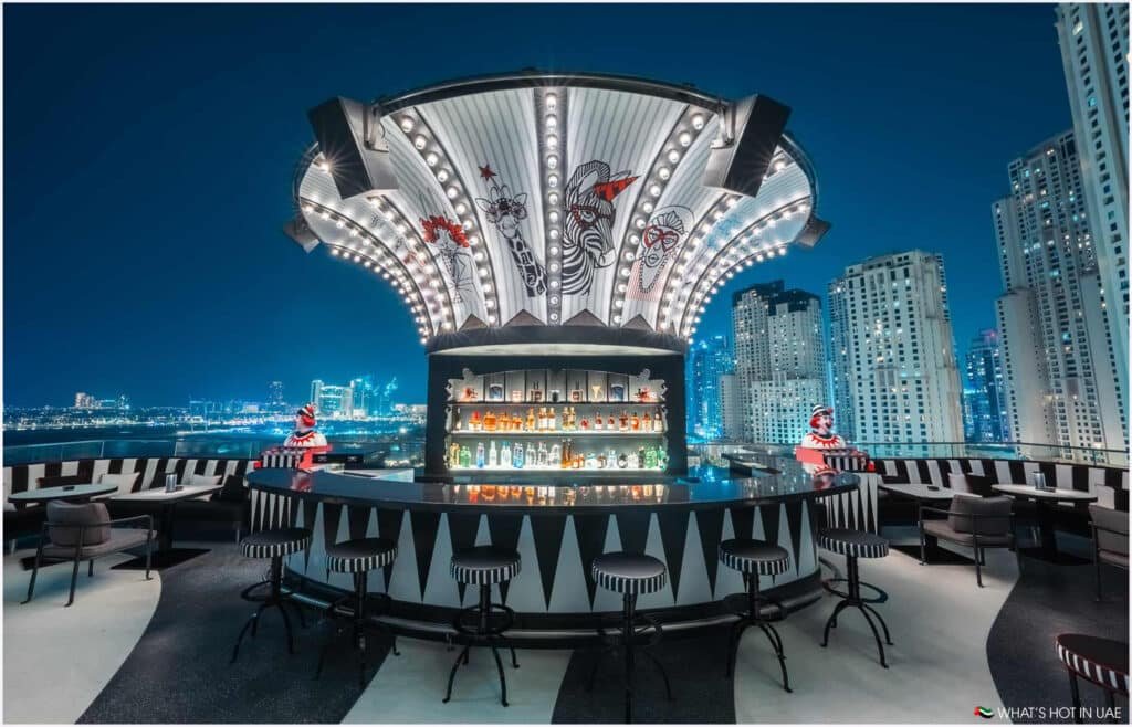 Rooftop bar with a circular counter adorned with striped bar stools and elaborate overhead lighting, featuring illustrative animal art. The setting overlooks a cityscape with tall buildings at night.