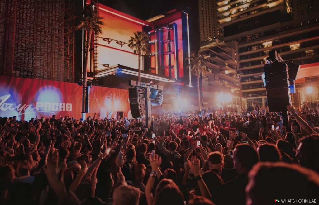 A large crowd enjoying a lively night event in front of an illuminated outdoor stage, surrounded by palm trees and buildings.
