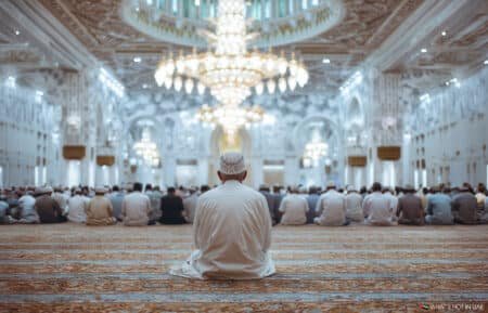 A man sits on the carpet in a large, ornate mosque, facing a congregation of people and an elaborate chandelier hanging from the intricately decorated ceiling.