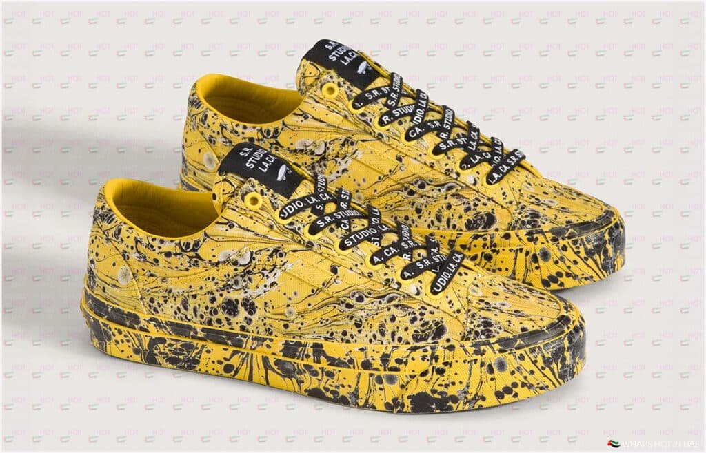 A pair of yellow sneakers with a black paint splatter design, black laces featuring white text, and a background with a subtle repeating pattern.