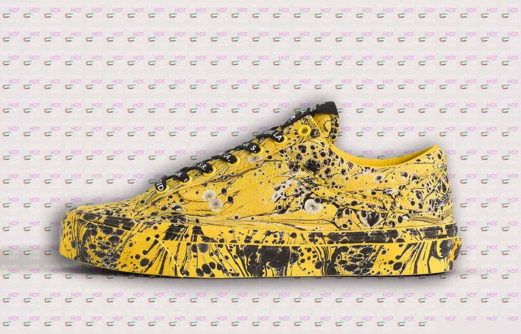 A yellow sneaker with black abstract splatter and swirl patterns on a patterned background featuring repeated text "What's Hot UAE" and small UAE flags.