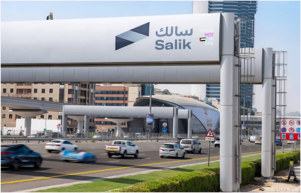 A highway in Dubai with cars in motion and an overhead Salik tollgate featuring the Salik logo and Arabic text. The background shows modern buildings and a metro station.