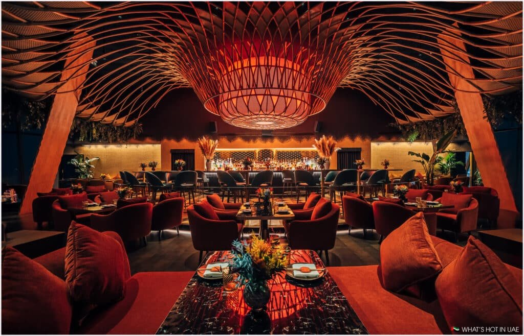 A luxurious restaurant interior with warm lighting, featuring plush red seating and elegant tables set with flowers. The ceiling is adorned with a large, intricate light fixture, and a well-stocked bar is visible at the back, surrounded by high chairs. The ambiance is cozy and inviting, enhanced by decorative plants and modern design elements.