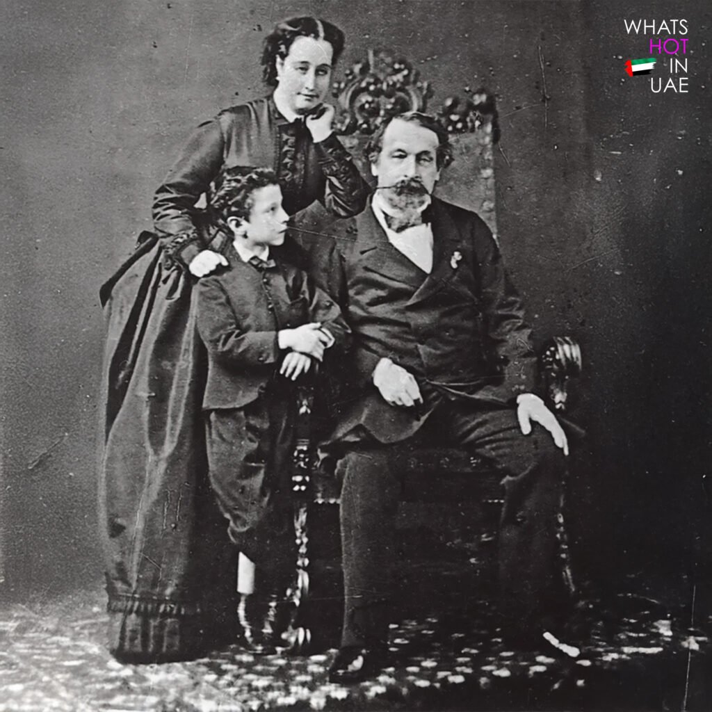 A vintage black and white photograph of three people from the late 19th century; an adult male seated in a formal suit, a woman standing behind with her hand on his shoulder, and a young boy leaning against him, all posed in front of a plain backdrop. This is Louis Vuitton and his family.