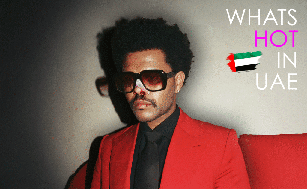 The Weeknd wearing sunglasses, a red jacket, and a black shirt with a bandaged nose stands against a plain background. The text 'What's Hot in UAE' with the UAE flag appears on the right side. Image made for the article about his 1 billion dollar deal.