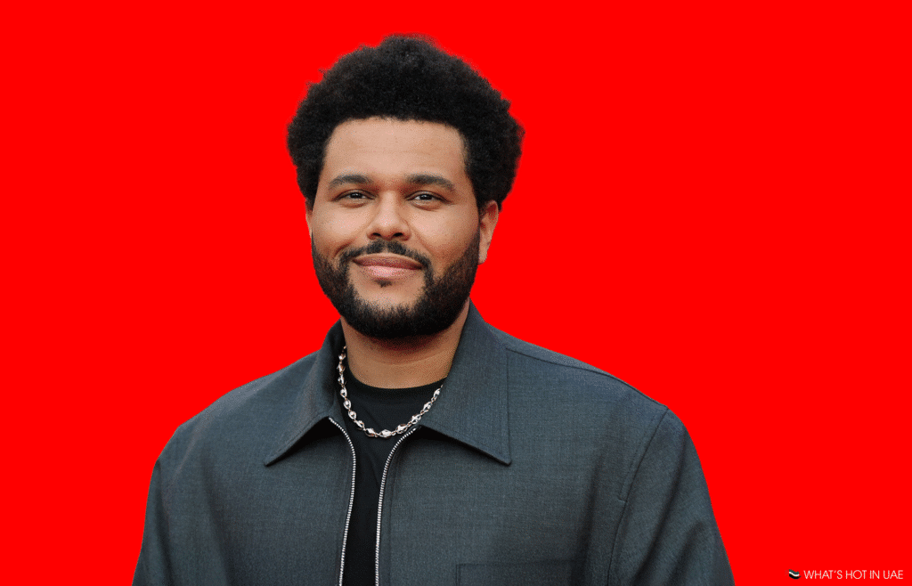 A man with curly black hair and a beard, wearing a dark jacket and a chain necklace, stands against a solid red background.