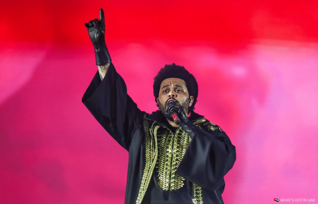 The Weeknd on stage wearing a black robe with gold embroidery, raising one hand and holding a microphone, set against a vibrant red background.