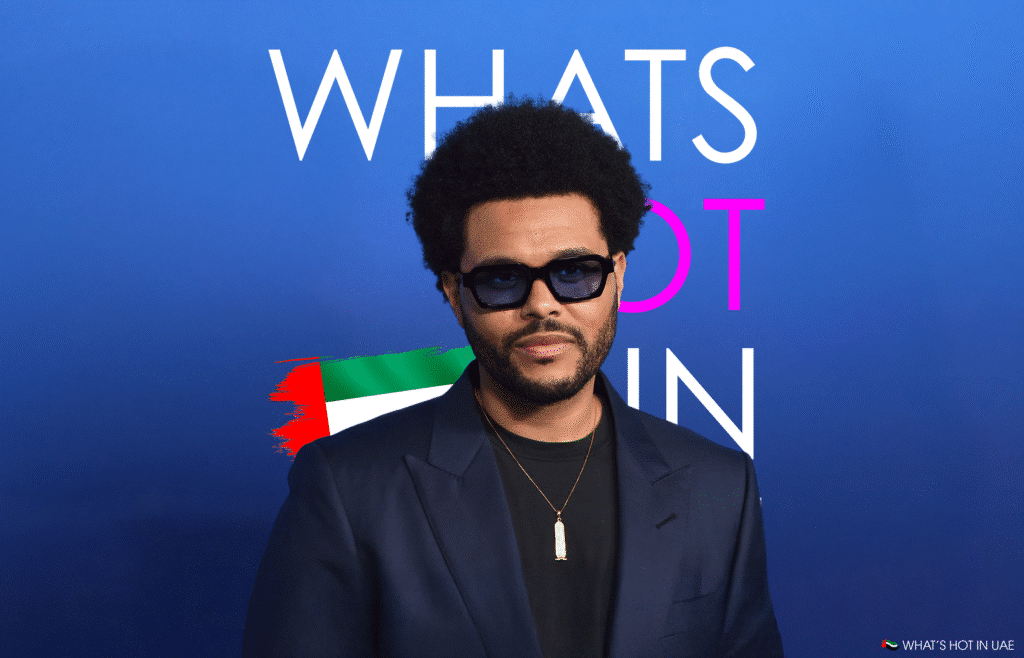The Weeknd wearing dark sunglasses and a dark suit stands in front of a blue backdrop with the text "WHAT'S HOT IN" partially visible behind him, and a small image of the UAE flag in the corner. His 1 billion dollar deal is the main focus of today.