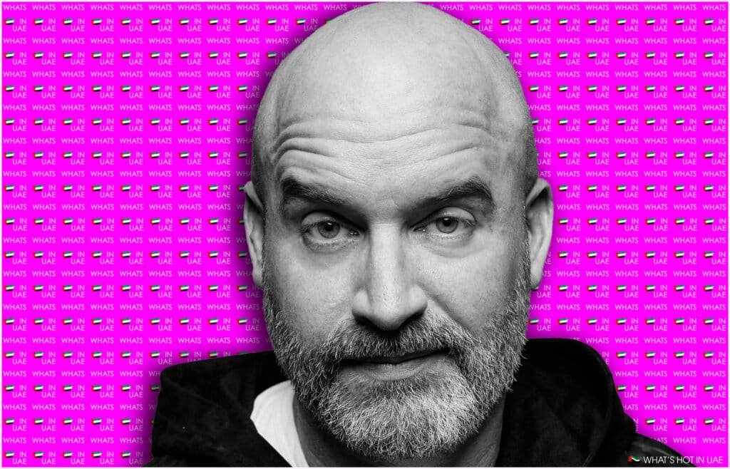 A black and white photo of Tom Segura with a beard against a vibrant pink background that repeats the phrase 'WHAT'S HOT IN UAE' alongside small UAE flags.
