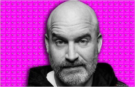 A black and white photo of Tom Segura with a beard against a vibrant pink background that repeats the phrase 'WHAT'S HOT IN UAE' alongside small UAE flags.
